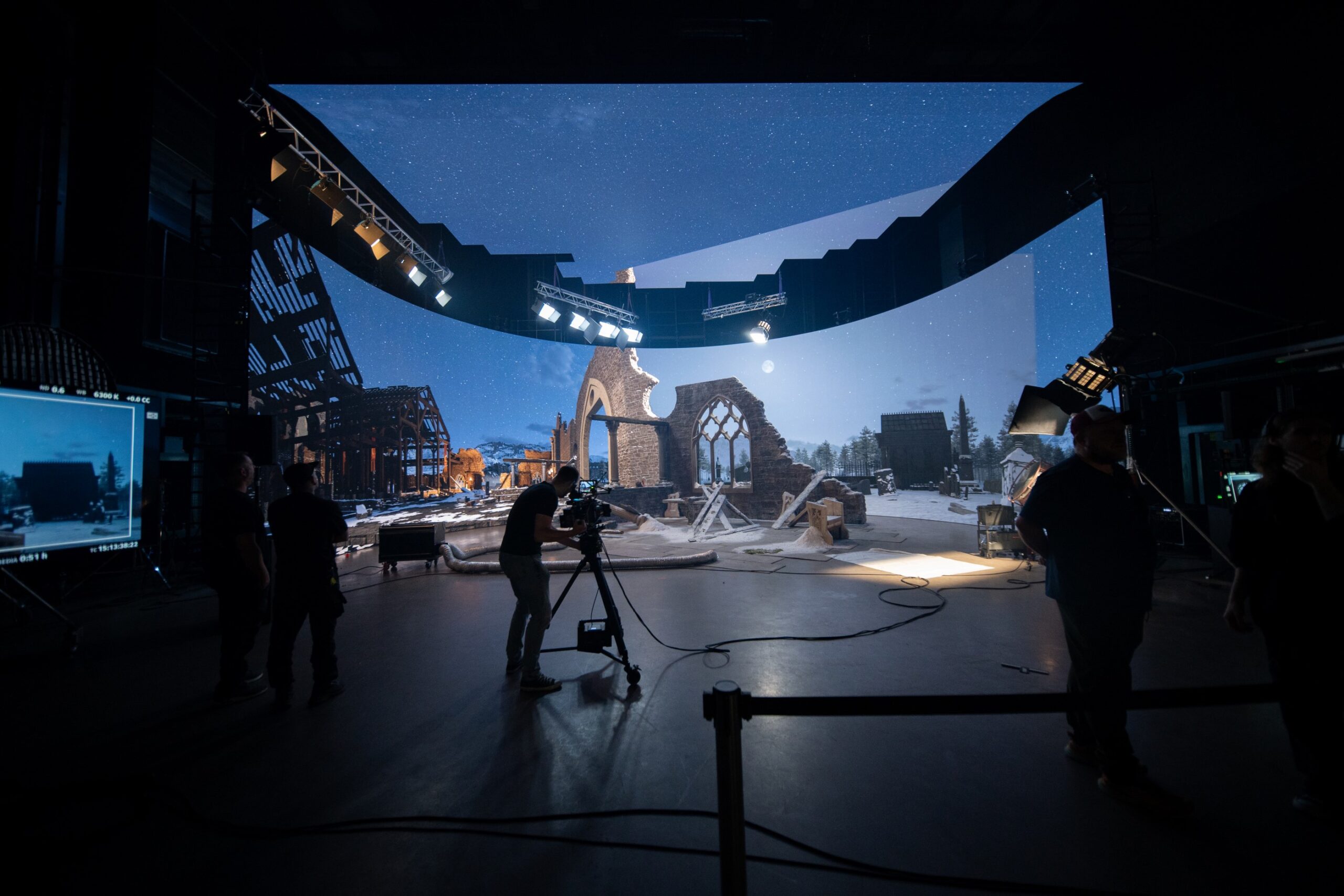 Film crew operating cameras on a virtual production set with a large curved LED screen displaying a night-time scene of ruined stone buildings.