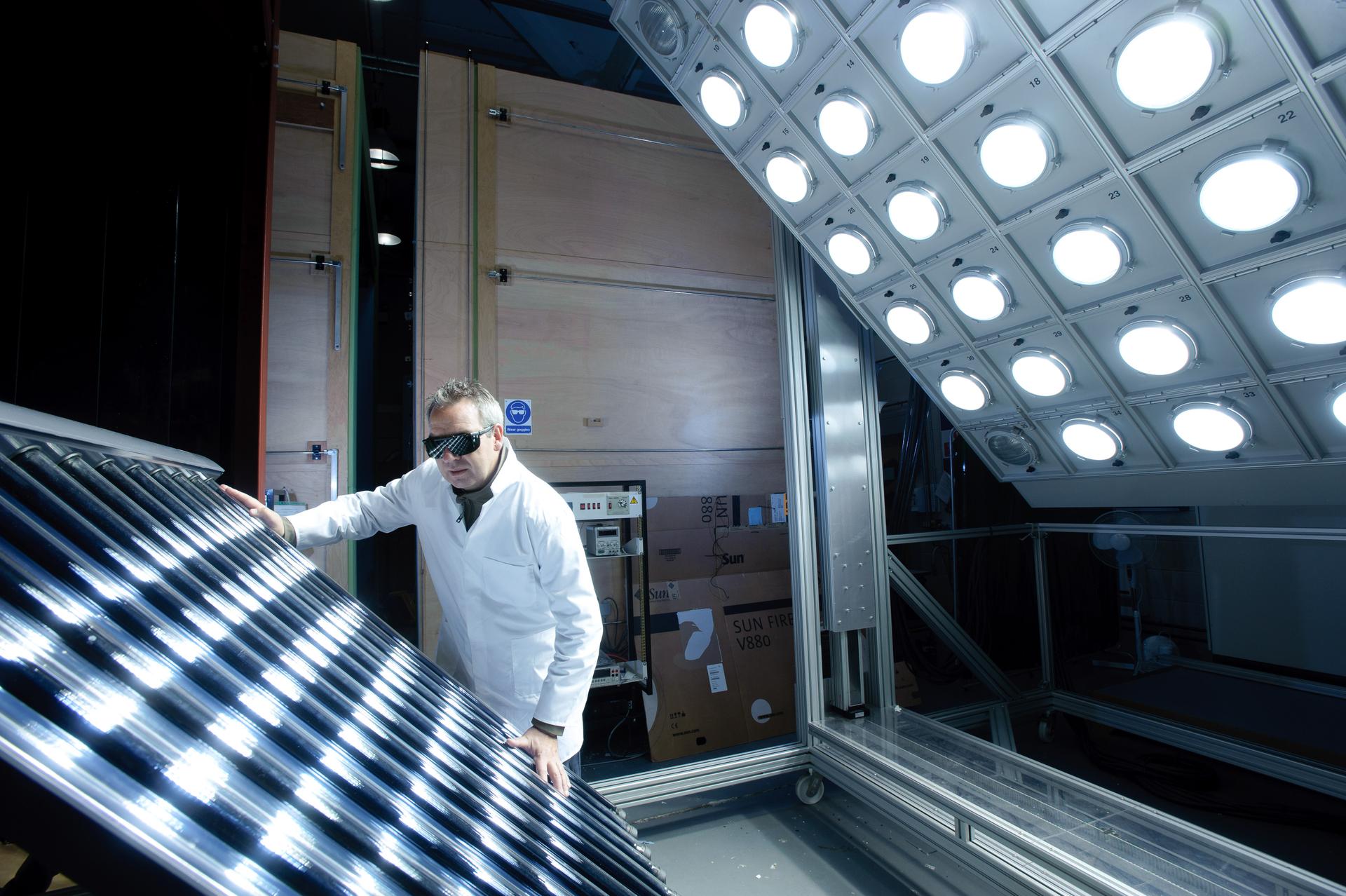 Researcher in a white lab coat and protective glasses operating solar energy testing equipment under an array of bright simulated sunlight lamps.