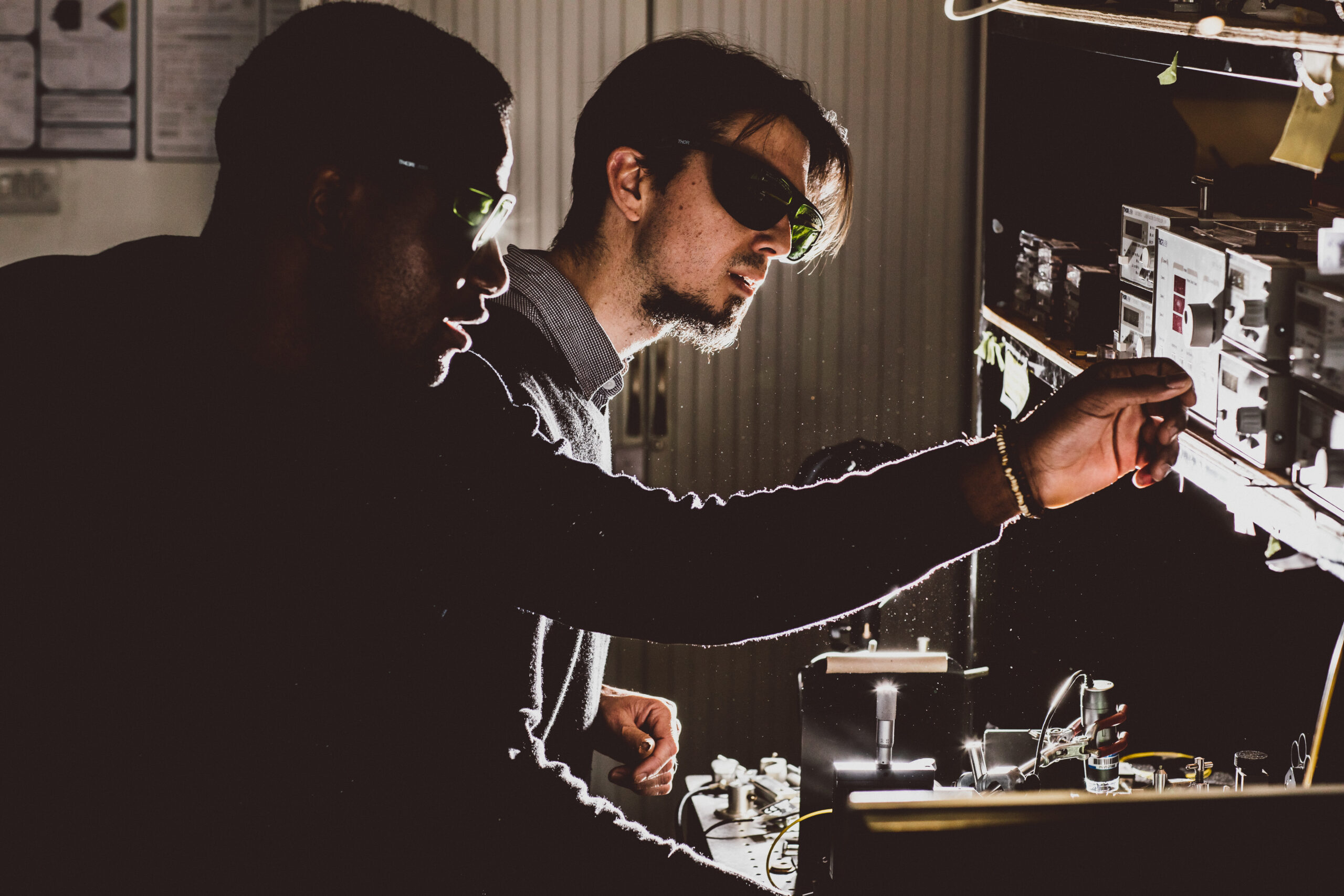 Two researchers wearing safety glasses work together in a dimly lit laboratory, adjusting equipment on a workbench illuminated by focused light.
